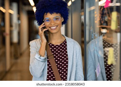 Portrait of happy biracial casual businesswoman with blue afro talking on phone in office corridor. Casual office, communication, business and work, unaltered. - Powered by Shutterstock