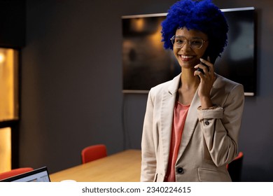 Portrait of happy biracial casual businesswoman with blue afro talking on smartphone in office. Casual office, communication, business and creative work, unaltered. - Powered by Shutterstock