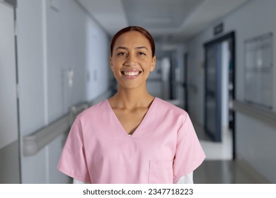 Portrait of happy biracial female doctor wearing scrubs in corridor at hospital. Hospital, medicine, healthcare and work, unaltered. - Powered by Shutterstock