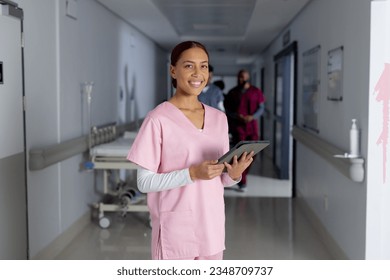 Portrait of happy biracial female doctor wearing scrubs holding tablet in corridor at hospital. Hospital, communication, medicine, healthcare and work, unaltered. - Powered by Shutterstock