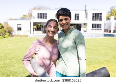 Portrait of happy biracial lesbian couple holding yoga mats embracing in sunny garden. Summer, gay, relationship, togetherness, yoga, fitness and healthy lifestyle, unaltered. - Powered by Shutterstock