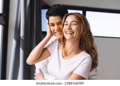 Portrait of happy biracial lesbian couple embracing in sun by window at home. Summer, gay, love, relationship, togetherness, domestic life and lifestyle, unaltered. - Powered by Shutterstock