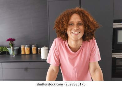 Portrait of happy biracial man with curly red hair leaning on counter in modern kitchen. Wellbeing, lifestyle and domestic life, unaltered. - Powered by Shutterstock