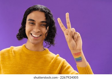 Portrait of happy biracial man showing v sign with lgbt flag on arm on purple background. Lgbt and pride concept. - Powered by Shutterstock