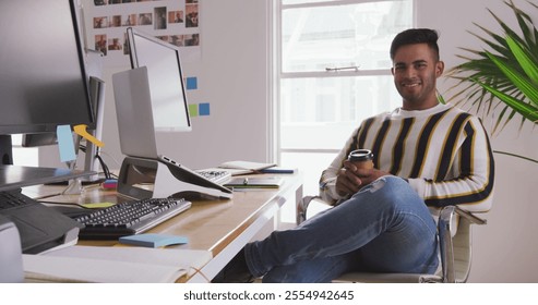 Portrait of a happy biracial man working in a creative office, using graphic tablet and laptop computer, holding a cup of take away coffee - Powered by Shutterstock