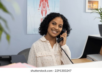 Portrait of happy biracial medical receptionist sitting at reception desk and talking on telephone. Hospital, medicine and healthcare.