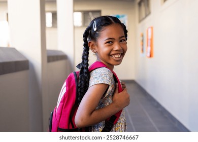 Portrait of happy biracial schoolgirl with school bag smiling in corridor at elementary school. Education, childhood, development, learning and school, unaltered. - Powered by Shutterstock