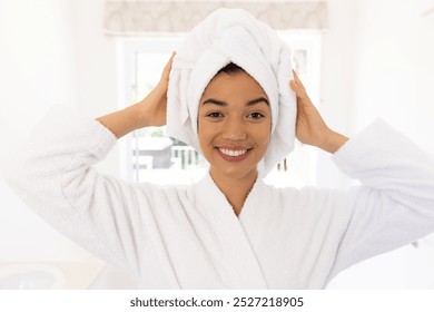 Portrait of happy biracial woman in bathrobe and with towel on her head in sunny bathroom. Lifestyle, self care and domestic life, unaltered. - Powered by Shutterstock