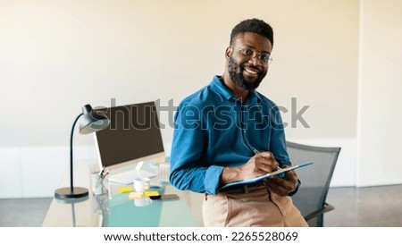 Portrait of happy black businessman standing in office, posing looking at camera holding clipboard and writing, taking notes, panorama, free space
