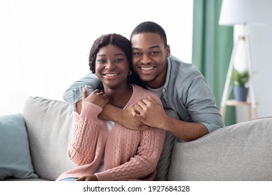 Portrait Of Happy Black Couple Or Husband And Wife Hugging At Home, Sitting On Couch In Living Room, Copy Space. Cheerful African American Man Embracing His Smiling Girlfriend From Behind