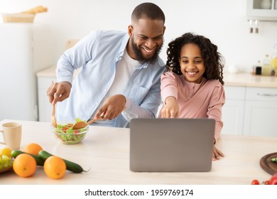 Portrait Of Happy Black Dad Cooking Dinner In Kitchen And Watching Food Video Blog On Laptop Or Reading Recipe Online Using Computer, Man Mixing Vegetable Salad, Daughter Showing And Pointing