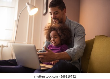 Portrait Of A Happy Black Dad Showing His Daughter How To Use Computer While She Is Sitting In His Lap Indoors.