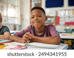 Portrait of happy black elementary boy studying in classroom while looking at camera. Close up face of little satisfied black boy writing in notebook using pencil. Proud schoolchild in primary school.