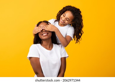 Portrait of happy black girl covering her excited mother eyes isolated on yellow orange studio background. Loving African American daughter making surprise for mom on holiday, playing Guess Who game - Powered by Shutterstock