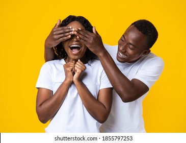 Portrait Of Happy Black Guy Covering His Excited Girlfriend Eyes Over Yellow Studio Background. Loving African American Husband Making Surprise For His Wife On Valentine Or Birthday