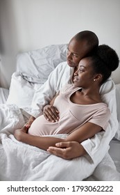 portrait of happy black married couple dreaming at home, handsome black guy hugs his pregnant wife, they look side and think