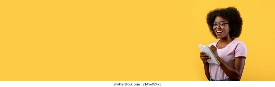 Portrait of happy black woman in eyeglasses taking notes in notebook and looking at camera, african american student writing down in notepad while posing on yellow background, wide horizontal banner - Shutterstock ID 2145692493