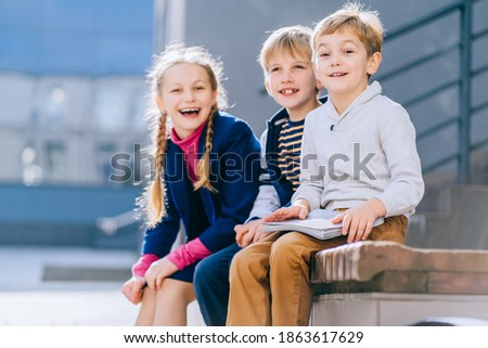 Portrait of happy blond little boy with other kids on blurred background.Three different age children with book sitting on the bench in the schoolyard at the day time.