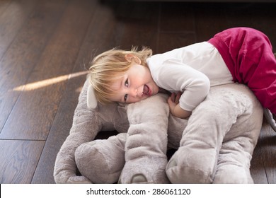 portrait of happy blonde caucasian baby nineteen month age looking at camera smiling  face embraced grey plush doll on brown wooden floor - Powered by Shutterstock