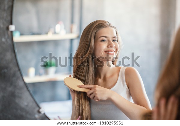 Portrait of happy blonde lady brushing her long\
hair near looking glass at\
bathroom