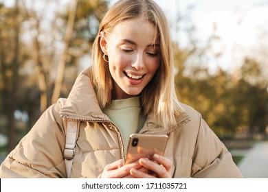 Portrait of happy blonde woman wearing jacket smiling and using cellphone while walking in park - Shutterstock ID 1711035721