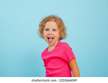 Portrait Of Happy Blue-eyed Little Girl With Curly Fair Hair Wearing Pink T-shirt, Looking, Showing Tongue, Grimacing Teasing On Blue Background. Studio. Advertisement, Emotions, Feelings, Copy Space.