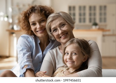 Portrait Of Happy Bonding Three Generations Family Posing In Modern Apartment. Sincere Smiling Young Woman Cuddling Elderly Mature Mother And Little Preschool Child Daughter, Looking At Camera.