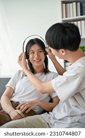 Portrait of a happy boyfriend putting on headphones for his girlfriend while spending time on the weekend at home together. - Shutterstock ID 2310744807