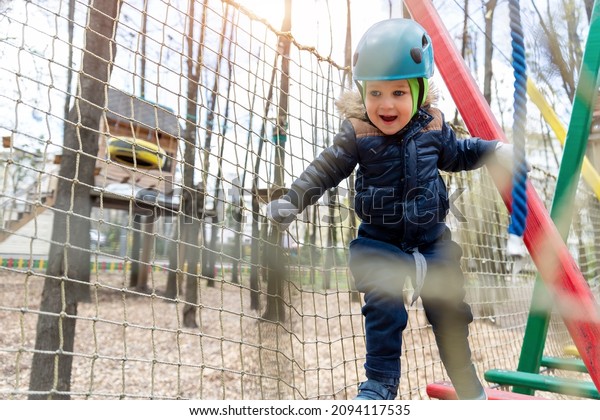 Portrait happy brave courage little toddler child\
boy wear safety equipment helmet enjoy passing obstacle course\
forest rope adventure park on cold winter day. Active outside\
leisure amusement camp