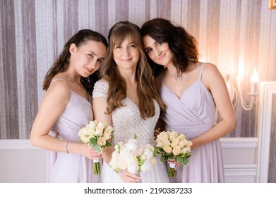 Portrait of happy bridesmaids bowing their heads on shoulders of pretty bride in wedding dress, girls posing with bouquets in hands, standing in apartment. Wedding haistyle and dresses concept.