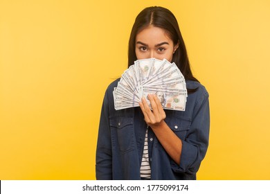 Portrait of happy brunette girl in denim shirt peeking out of dollar banknotes with playful happy look, showing money, big profit, financial savings. indoor studio shot isolated on yellow background