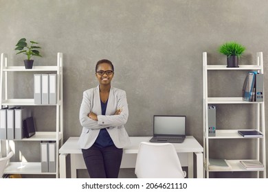 Portrait Of Happy Business Lady At Work. Beautiful Young Black Woman In Suit And Glasses Leaning On Desk While Standing Arms Crossed In Modern Office Workplace Interior