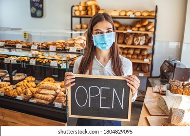 Portrait Of A Happy Business Owner With An Open Sign On The Bakery Shop And Smiling - Food And Drinks Concepts. Woman Working At A Bakery Wearing A Facemask To Avoid The Coronavirus