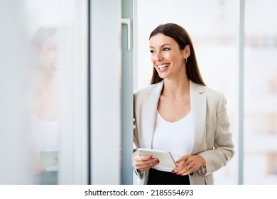 Portrait Of Happy Businesswoman Wearing Blazer And Using Digital Tablet While Standing By The Widow At The Office. 