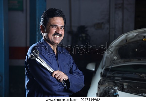 Portrait of a happy car mechanic in moustache\
standing beside a car in service station. Car Specialist is holding\
car repairing tools. Professional repairman is smiling in modern\
clean work workshop.