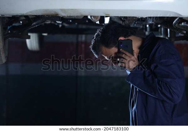 Portrait of a happy car mechanic in moustache\
talking on phone while examining the lifted car. Car specialist is\
holding car repairing tools. Repairman wearing a blue mechanic\'s\
uniform.