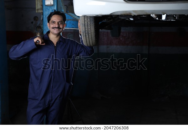 Portrait of happy car mechanic in moustache\
standing beside a lifted car in service station. Car Specialist is\
holding car repairing tools. Professional repairman is wearing\
mechanic uniform in\
blue.