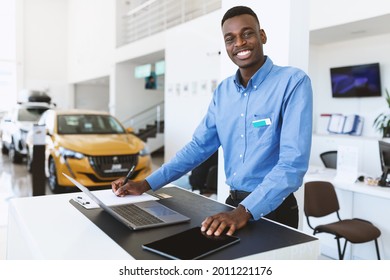 Portrait of happy car salesman standing at work desk, smiling at camera, using laptop and touch pad in auto dealership. Positive black automobile manager posing at workplace in showroom store