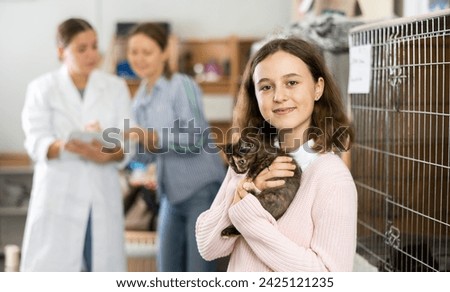 Portrait of happy caring preteen girl helping in animal shelter, standing near cages with frightened motley kitten in arms ..