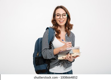 Portrait of a happy casual girl student with backpack writing in a notepad while standing with books isolated over white background - Powered by Shutterstock