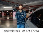 Portrait of happy casual man leaning on his car with arms crossed and smiling at the camera in underground parking lot.