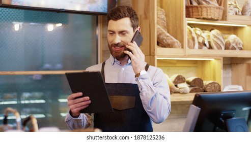 Portrait of happy Caucasian bearded handsome male baker standing in own bakery, tapping on tablet device and speaking on smartphone. Male worker talking on mobile phone with client. Bakehouse concept - Powered by Shutterstock