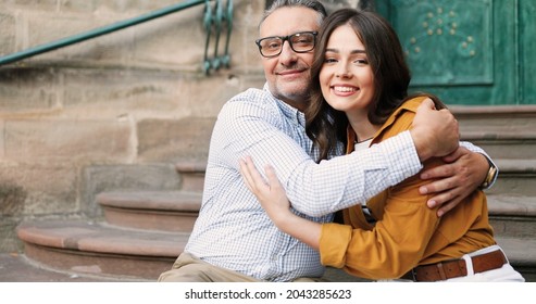 Portrait of happy Caucasian father in glasses hugging pretty adult daughter outdoor while sitting on steps amd smiling to camera. Beautiful cheerful young girl smiling in hugs of dad. Fatherhood love.