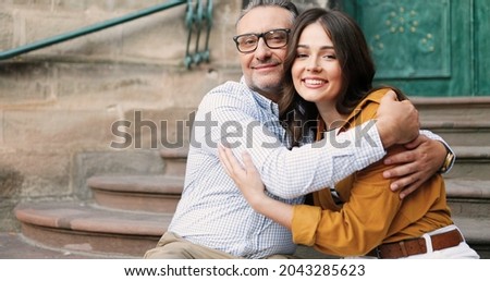 Portrait of happy Caucasian father in glasses hugging pretty adult daughter outdoor while sitting on steps amd smiling to camera. Beautiful cheerful young girl smiling in hugs of dad. Fatherhood love.