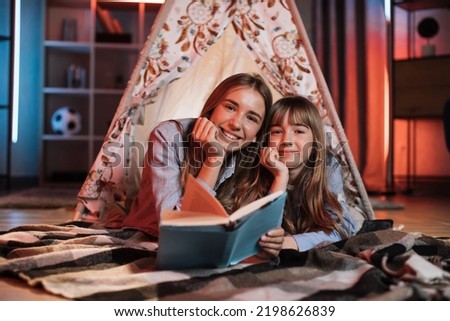 Portrait of happy caucasian loving sisters smiling and looking at camera inside wigwam. Beautiful girls and open book lying near on floor.
