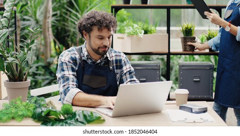 Portrait of happy Caucasian male and female florists in aprons working at flower shop and doing inventory. Man typing on laptop at desk in greenhouse. Woman tapping on tablet. Family business concept - Powered by Shutterstock