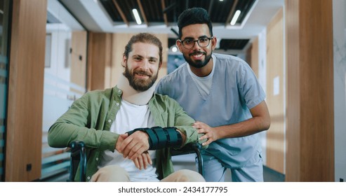 Portrait of happy Caucasian male patient injured in wheelchair recovering in modern hospital with professional Indian doctor in uniform. Handsome men looking at camera and smiling in hospital. - Powered by Shutterstock