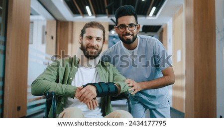 Portrait of happy Caucasian male patient injured in wheelchair recovering in modern hospital with professional Indian doctor in uniform. Handsome men looking at camera and smiling in hospital.