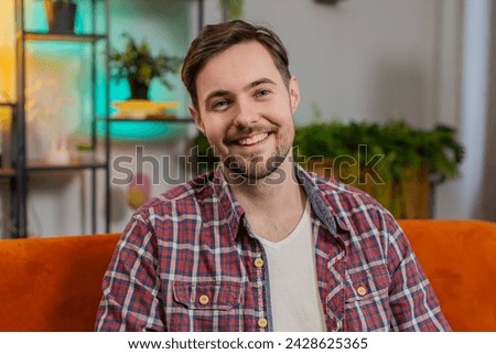 Portrait of happy Caucasian man sitting on sofa looking at camera and smiling at home. Young guy in plaid shirt having attractive appearance enjoying leisure time relaxing in living room in apartment