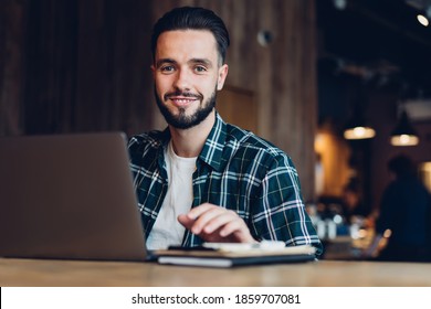 Portrait of happy Caucasian man smiling at camera while doing web project via laptop application, successful millennial freelancer 20 years old posing at table desktop with modern netbook computer